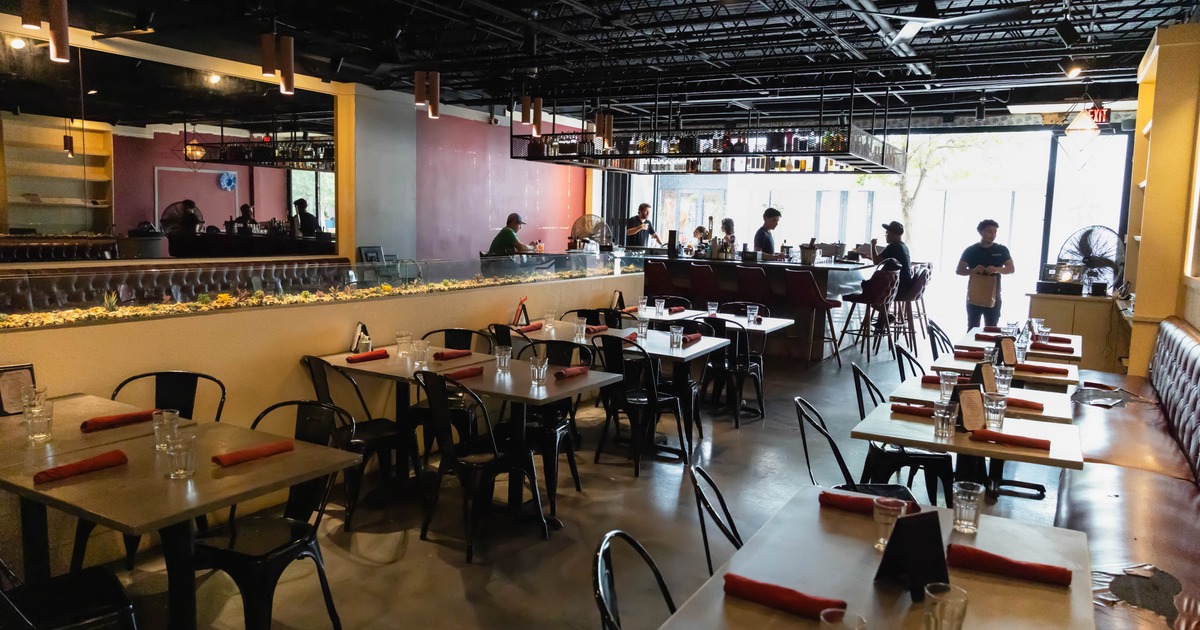 Interior of a modern restaurant with tables set with red napkins, a bar, and large windows