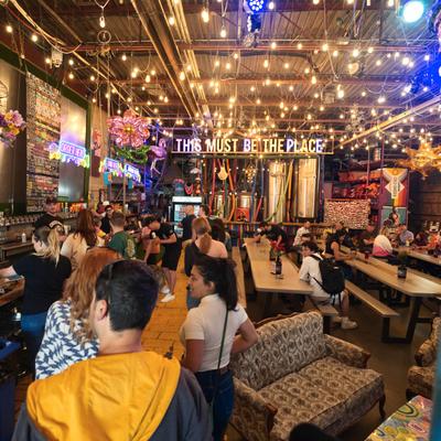 Crowded brewery taproom with communal seating, string lights, and neon signs.