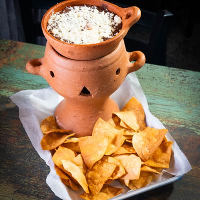 Refried beans topped with fresh cheese in a warm clay pot served with tortilla chips.