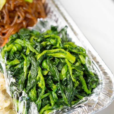 Close up, spinach placed in a foil container, next to brown stir-fried noodles.