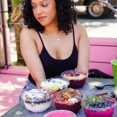 Outdoors, a person sitting by a table with assorted acai bowls.