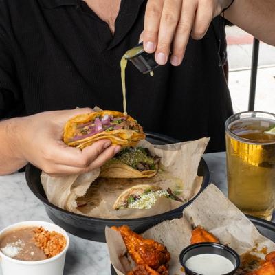 A person at the restaurant enjoying tacos and chicken wings with a glass of beer.