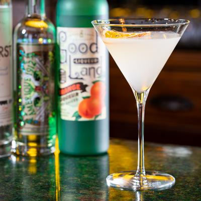 Citrus martini in tall glass beside colorful liquor bottles on bar counter.