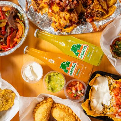 Various food dishes and bottles of Mexican soda, with sides of guacamole, sour cream, and salsa.
