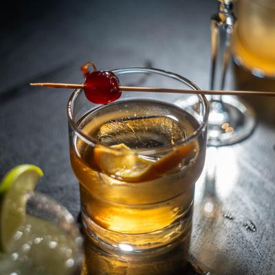 Whiskey drink with citrus peel and cherry garnish.