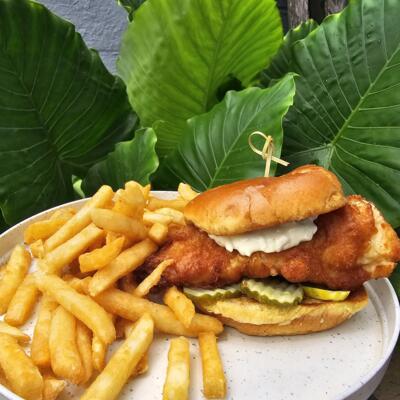 Beer-battered fish sandwich with pickles, served with fries against vibrant green leaves.
