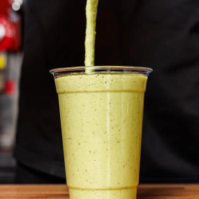 Green fruit smoothie being poured  in a clear plastic cup on a wooden counter.
