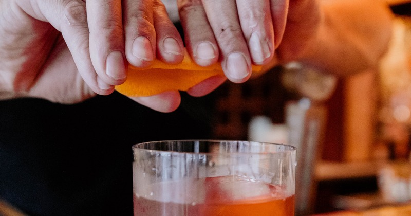 Bartender squeezing orange peel over a whiskey drink.