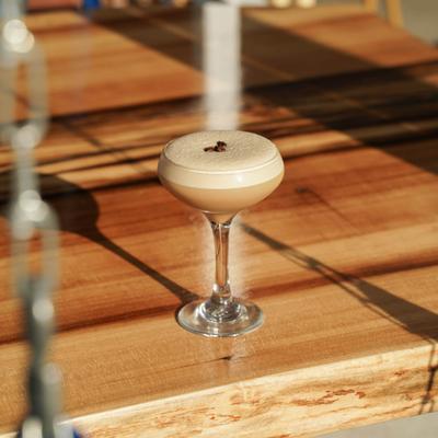 An espresso martini rests on a sunny wooden outdoor table.