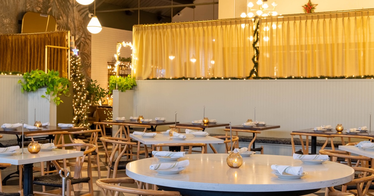 A warmly lit interior with tables set with plates, napkins and candle lanterns; wooden chairs