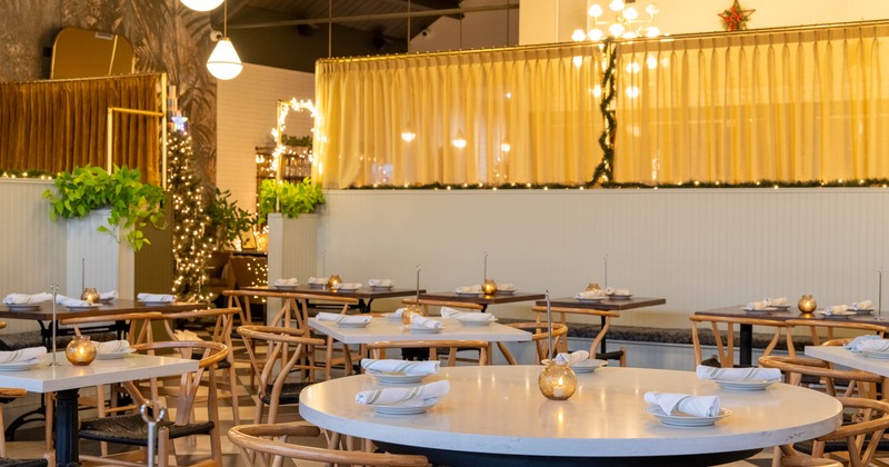 A warmly lit interior with tables set with plates, napkins and candle lanterns; wooden chairs