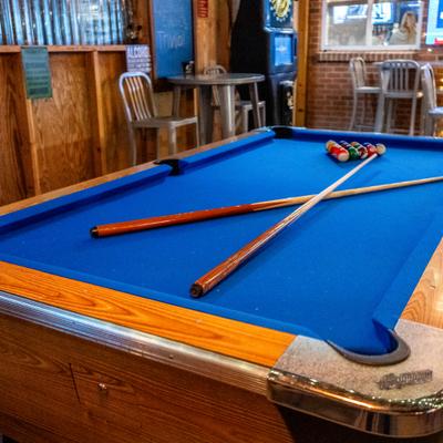 Bar interior, a pool table with blue felt surface, two pool cues, and a rack of billiard balls.