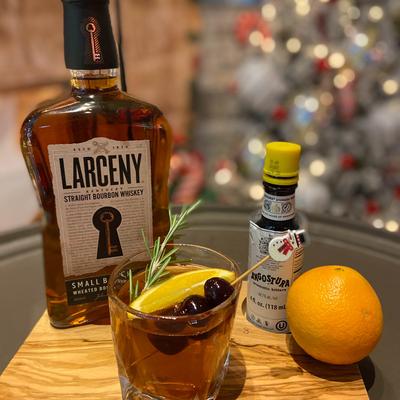 A bourbon cocktail garnished with citrus, cherries, and rosemary beside a full bottle.
