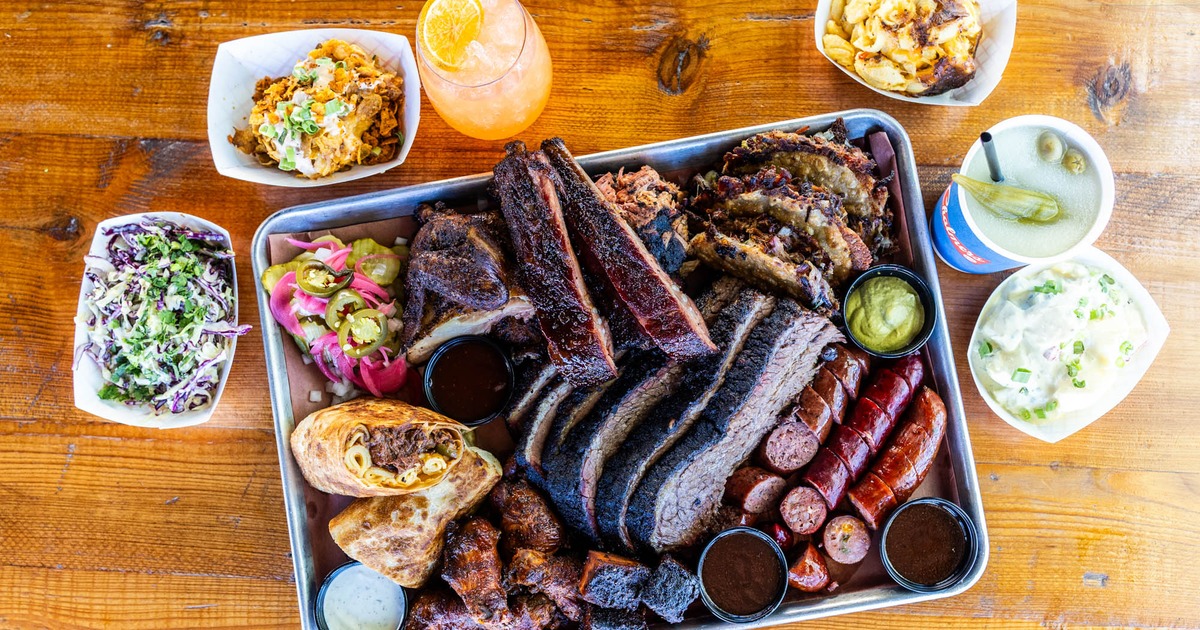 A large tray of barbecue from Rollin Smoke BBQ, surrounded by side dishes, and drinks