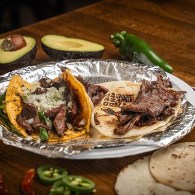 Carne asada taco plate on a table with halved avocado, lime wedges, raw jalapenos.