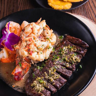 Grilled steak and lobster tail on a black plate, garnished with herbs and vibrant purple flowers.