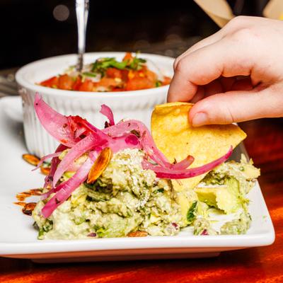 Hand dipping chips into a guacamole topped with pickled red onions and nuts.