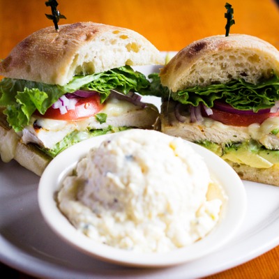 Chicken sandwich on a cibatta roll with lettuce, tomato and avocado inside. Plated next to a serving of potato salad