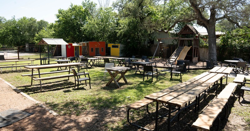 Outdoor seating area, picnic tables, chairs, a tree, colorful structures, and a playset with slide