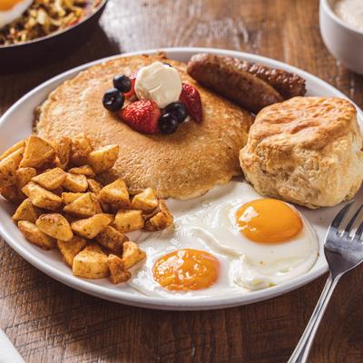 Pancakes topped with berries, accompanied by fried eggs, home fries, sausages, and a biscuit