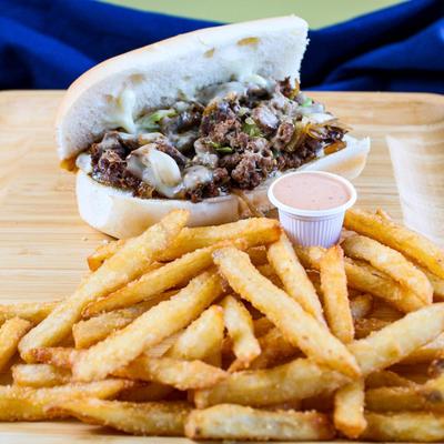 Philly cheesesteak served on a wooden plate with fries and a dipping sauce.
