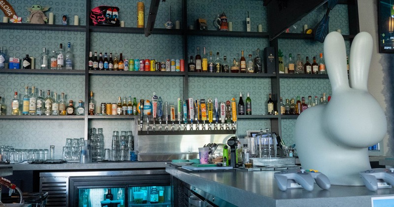 A bar interior with liquor bottles on shelves, colorful beer taps, a white rabbit sculpture
