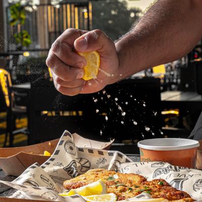 Hand squeezing a fresh lemon over a schnitzel served in patterned paper.