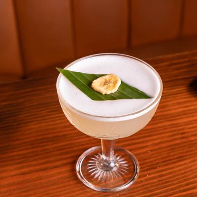 Frothy cocktail with a banana slice and a leaf garnish on a wooden table.