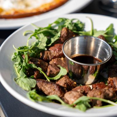 Steak bites served on a bed of arugula with a cup of sauce.