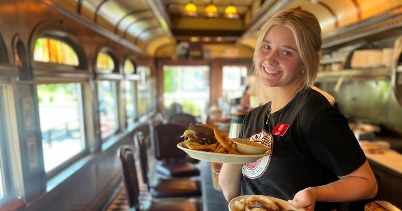 A smiling waitperson holds two plates of food inside a diner