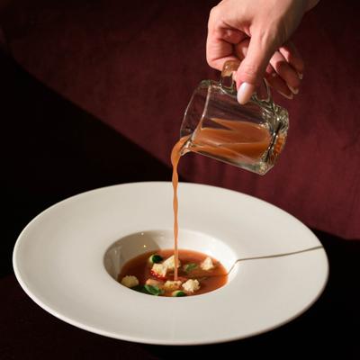 Soup being poured from a small glass pitcher into a wide-rimmed white bowl.