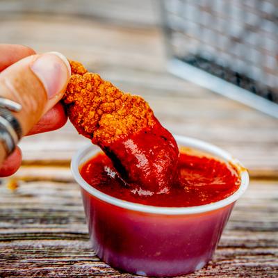 A crispy popcorn chicken piece being dipped into a small cup of red sauce.