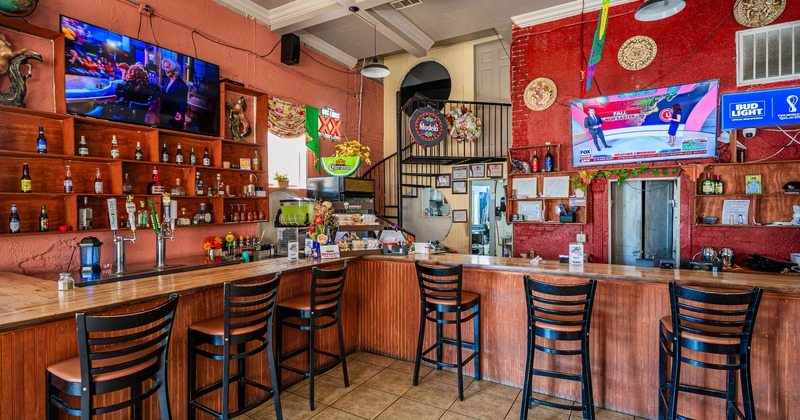 Wooden bar counter with high chairs and overhead TVs in a colorful Mexican restaurant