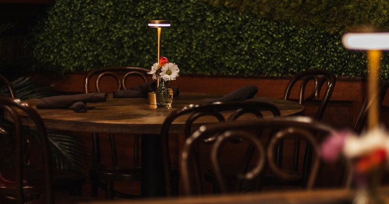Round wooden table with black napkin roll-ups, flowers in a vase, and a lamp