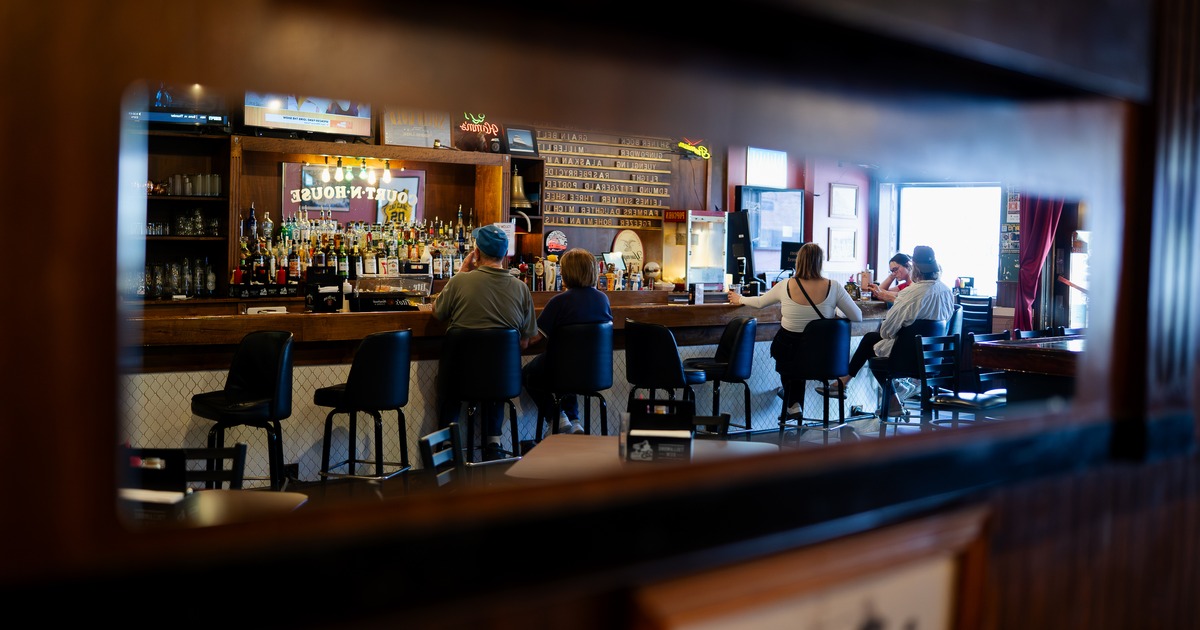 Bar, chairs, guests sitting, drinking, drinks, drink rack behind, photo in mirror on wall across