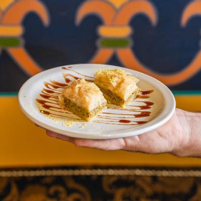 A hand holding a white plate with two pieces of baklava, in front of an ornate wall.