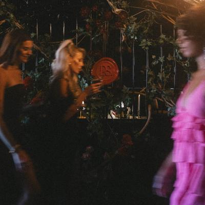 Three models walk past a lush backdrop of ivy and red flowers in a dimly lit setting.