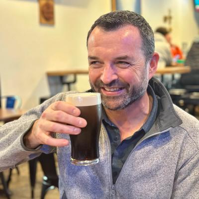A smiling guest holds a dark beer with a frothy head.