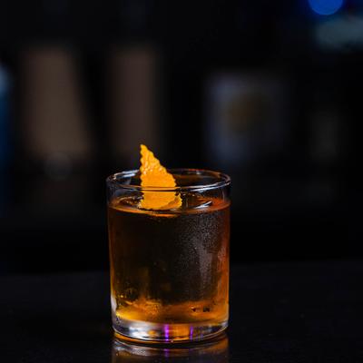 A glass of amber drink, topped with an orange peel, sits on a reflective dark surface.