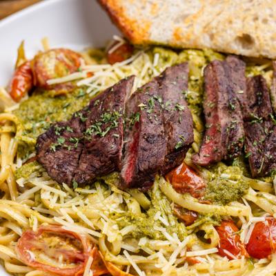Plate of pesto pasta with sliced steak, cherry tomatoes and toasted bread.