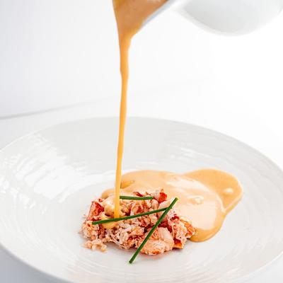 Pouring creamy orange sauce over chopped lobster garnished with green chives on a white plate.