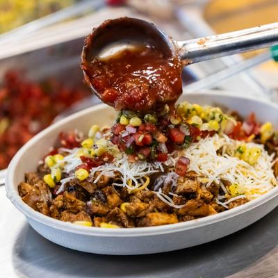 A ladle pouring salsa over a bowl of rice, chicken, and cheese.