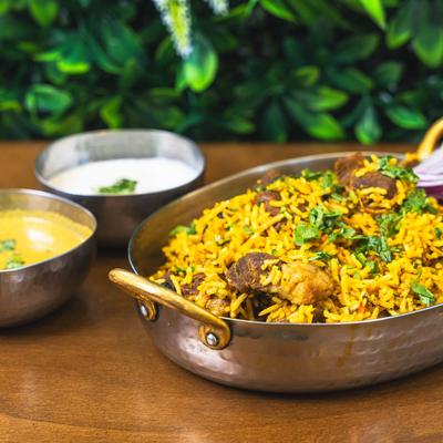 Lamb biryani served in a copper bowl with raita and curry on the side.