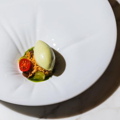 A scoop of light green sorbet sits on a white plate with a cherry tomato, a grape, and green sauce.