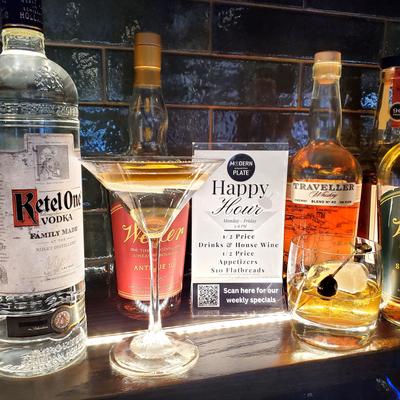 Bar shelf featuring a bottles of spirits, two cocktails, and a promotional happy hour sign.