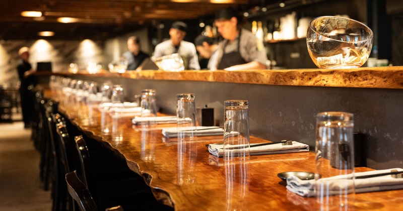 Warmly lit restaurant bar with a long wooden counter, table settings, glassware
