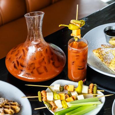 Decanter and individual glass filled with a Bloody Mary, alongside garnishes and meals d.