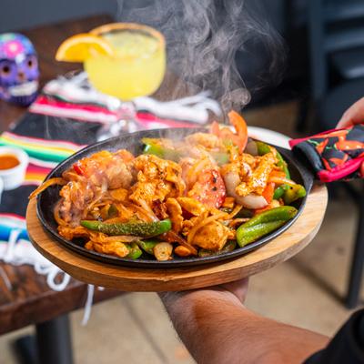 A person holding a skillet of sizzling chicken fajitas with cocktails in the background.