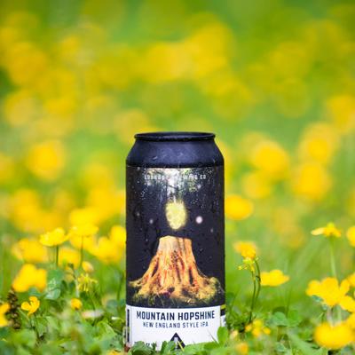 Can of Mountain Hopshine New England Style IPA stands on grass, surrounded by yellow flowers.