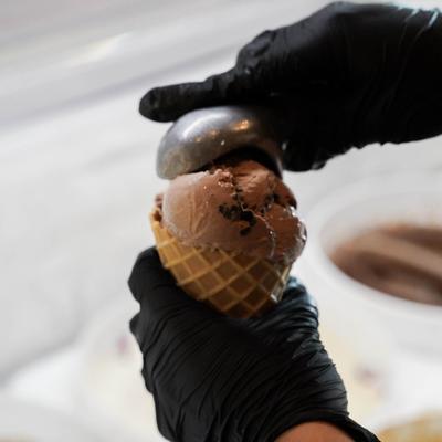 A gloved hand holding a chocolate ice cream cone, with chunks of chocolate & a waffle cone.